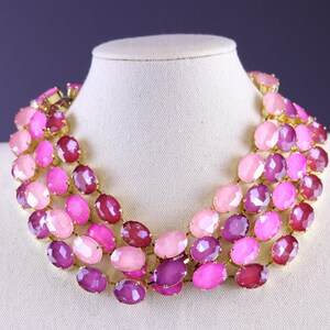 Può includere: Collana a tre fili con pietre preziose ovali rosa e viola. La collana è esposta su un busto di manichino bianco su sfondo viola scuro. Le pietre preziose sono in diverse tonalità di rosa e viola.