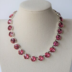 Può includere: Una collana color argento con grandi cristalli rotondi color rubino. I cristalli sono incastonati in castoni individuali e uniformemente distanziati. La collana è esposta su una testa di manichino su sfondo bianco.