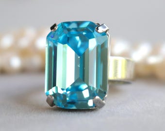 May include: A silver ring featuring a large, rectangular, light blue gemstone. The gemstone is faceted, reflecting light, and is set with four silver prongs. The ring's band is a simple, polished silver.
