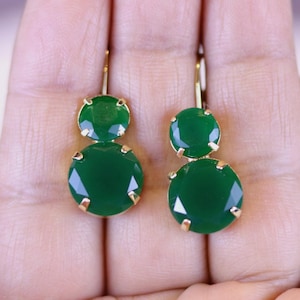 May include: A pair of gold-toned drop earrings featuring two round, faceted, emerald-green gemstones. The earrings have a lever-back closure and are displayed against a neutral background.
