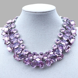 May include: A statement necklace featuring a cascade of large, faceted, lavender-colored crystals. The crystals are set in a silver-toned metal and arranged in a bib style, creating a glamorous and eye-catching design. The necklace is displayed on a white mannequin.