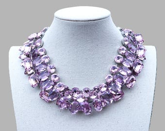 May include: A striking necklace with large, lavender-coloured crystals. The crystals are set in a silver-toned metal and arranged in a bib style. The necklace is displayed on a white mannequin. The crystals are arranged to create a glamorous design.