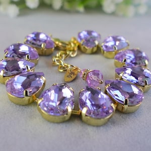 May include: A gold-toned bracelet featuring large, pear-shaped, light purple crystals. The crystals are set in gold bezels and linked together with a gold chain. A small gold charm is attached to the chain.