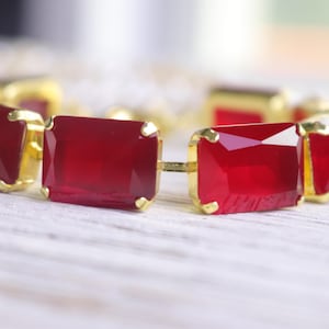 May include: A gold-toned bracelet featuring rectangular, faceted, deep red gemstones. The gemstones are set in gold-colored metal links, creating a bold and elegant accessory. The bracelet is displayed on a light-colored, textured surface.