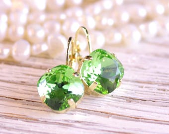 May include: A pair of gold-colored leverback earrings with large, faceted, light green gemstones. The earrings are set against a blurred background of white pearls and a light-colored wooden surface.