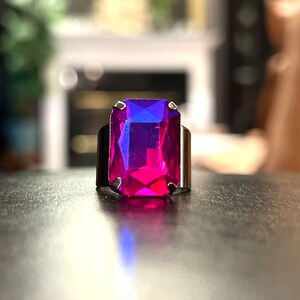 May include: A silver ring with a large, rectangular, faceted gemstone. The gemstone is a deep pink color with a blue hue.