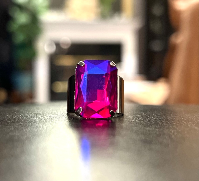 May include: A silver ring with a large, rectangular, faceted gemstone. The gemstone is a deep pink color with a blue hue.