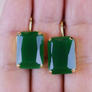 May include: A pair of emerald green earrings with a rectangular cut. The earrings have a gold-colored setting and a lever back closure. The gemstones are a deep, rich green color, and the earrings are displayed against a neutral background.