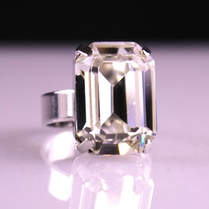 May include: A silver ring featuring a large, rectangular, clear gemstone with multiple facets. The gemstone is set in a silver band, creating a striking and elegant piece of jewelry. The ring is displayed on a reflective surface.