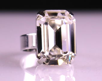 May include: A silver ring featuring a large, rectangular, clear gemstone with multiple facets. The gemstone is set in a silver band, creating a striking and elegant piece of jewelry. The ring is displayed on a reflective surface.
