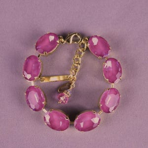 Può includere: Un bracciale dorato con grandi pietre preziose ovali color magenta. Il bracciale ha un'estensione a catena per la regolazione della taglia e una chiusura a moschettone. Le pietre sono incastonate in metallo dorato, creando un look classico ed elegante.