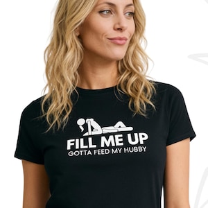 Op de afbeelding: Een zwart T-shirt met korte mouwen voorzien van een witte afbeelding van een liggend persoon en de tekst "FILL ME UP" gevolgd door "GOTTA FEED MY HUBBY" in witte hoofdletters. Het shirt heeft een ronde hals en een standaard pasvorm.