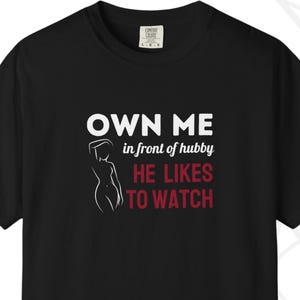 May include: Black t-shirt with white text that reads "OWN ME in front of hubby HE LIKES TO WATCH" and a line drawing of a woman. The shirt is from Bold Swing Apparel.
