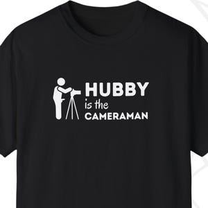 May include: Black t-shirt with white text that reads "Hubby is the cameraman" and a graphic of a person holding a camera on a tripod.