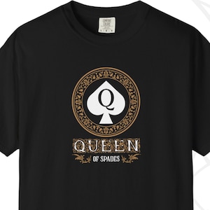 May include: Black t-shirt featuring a gold and white design of the Queen of Spades playing card. The design includes the word "QUEEN" above "OF SPADES" in a decorative font. The shirt is made of a soft material.