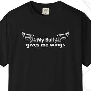May include: Black t-shirt with the text "My Bull gives me wings" in white, accompanied by a pair of stylized wings. The shirt is a short-sleeved crew neck style.