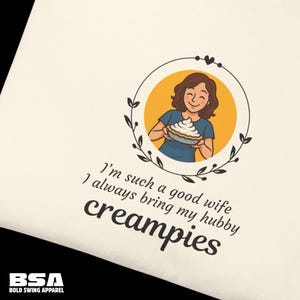 May include: Cream-coloured t-shirt featuring a graphic of a woman holding a pie, encircled by a floral wreath. Text reads "I'm such a good wife, I always bring my hubby creampies."