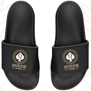 May include: Pair of black slide sandals featuring a Queen of Spades design. The sandals have a black base with a wide strap displaying a gold and white emblem. The text "QUEEN OF SPADES" is printed below the emblem.