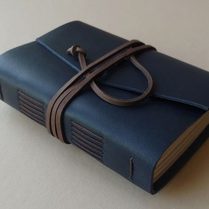 May include: A navy blue leather journal with a brown leather strap closure. The journal has stitched edges and a flap closure. The pages are visible and appear to be made of a natural paper. The journal is approximately 5 inches long and 4 inches wide.