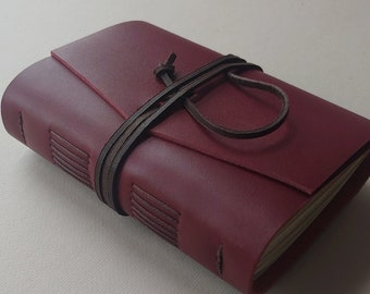 May include: A burgundy leather journal with a wraparound brown leather tie closure. The journal has a stitched spine and a flap that covers the top. The pages are visible at the top edge of the journal.