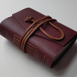 May include: A small, rectangular journal bound in deep burgundy textured leather. The cover is secured by a thin, light brown leather cord that wraps around the center and ties in a knot. The spine features visible stitching with light brown thread.