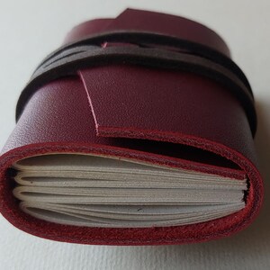 May include: A burgundy leather-bound journal with a black leather strap closure. The journal is closed and the strap is wrapped around it.