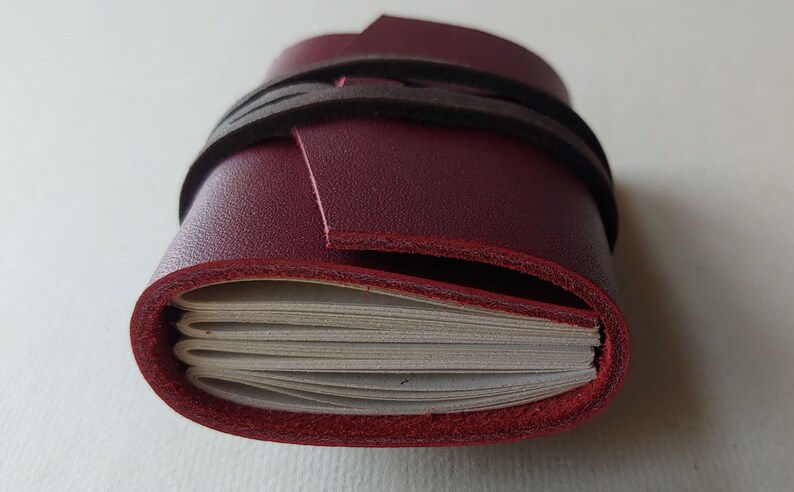 May include: A burgundy leather-bound journal with a black leather strap closure. The journal is closed and the strap is wrapped around it.