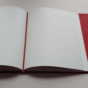 May include: An open journal with blank white pages and a red cover. The journal is bound with a red spine and a red flap that closes with a black string. The pages are clean and ready for writing or sketching.