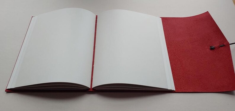 May include: An open journal with blank white pages and a red cover. The journal is bound with a red spine and a red flap that closes with a black string. The pages are clean and ready for writing or sketching.