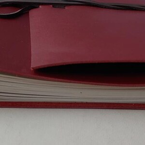 May include: A burgundy leather journal with a wrap-around closure. The journal's pages are visible, and the cover has a pocket. The leather is a deep red color, and the closure is a dark brown leather strap.