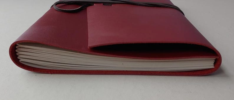 May include: A burgundy leather journal with a wrap-around closure. The journal's pages are visible, and the cover has a pocket. The leather is a deep red color, and the closure is a dark brown leather strap.