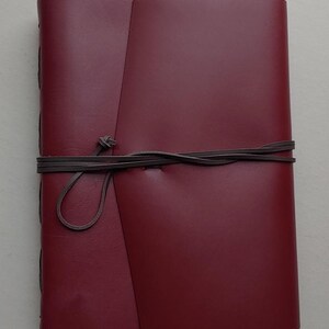 May include: A burgundy leather journal with a wrap-around closure. The journal is bound with dark brown leather cord. The cover is a solid burgundy color. The journal is closed with a leather cord that is tied in a knot.