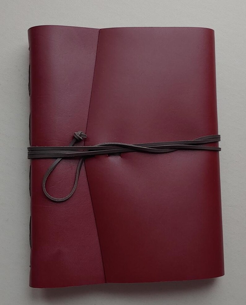 May include: A burgundy leather journal with a wrap-around closure. The journal is bound with dark brown leather cord. The cover is a solid burgundy color. The journal is closed with a leather cord that is tied in a knot.