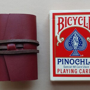 May include: A burgundy leather card case with brown leather straps. The case is next to a deck of Bicycle brand playing cards with a red background and a black spade suit. The card deck is labelled "PINOCHLE Special 48 Card Deck PLAYING CARDS".