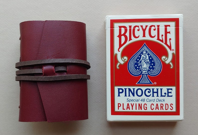 May include: A burgundy leather card case with brown leather straps. The case is next to a deck of Bicycle brand playing cards with a red background and a black spade suit. The card deck is labelled "PINOCHLE Special 48 Card Deck PLAYING CARDS".