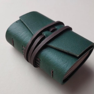 May include: A small, rectangular, dark green leather journal secured with a dark brown leather wrap-around cord. The journal features visible stitching along the spine and edges, with the cover folded over to protect the inner pages.