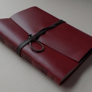 May include: A burgundy leather journal with a dark brown leather strap closure. The journal's cover is smooth, with embossed detailing along the spine. The strap wraps around the journal, securing the cover. A stylish notebook for writing.