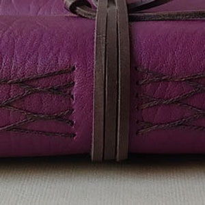 Puede incluir: Un diario de cuero de color morado oscuro con costuras decorativas y una correa de cuero marr&oacute;n.