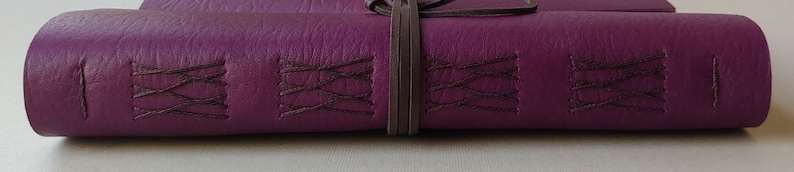Puede incluir: Un diario de cuero de color morado oscuro con costuras decorativas y una correa de cuero marr&oacute;n.
