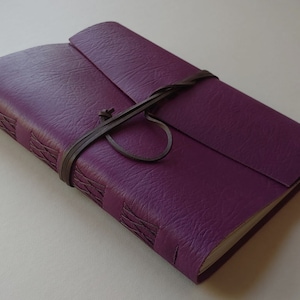 Puede incluir: Un diario encuadernado en cuero morado con una correa de cuero marr&oacute;n y un nudo decorativo. El diario tiene una superficie texturizada y est&aacute; cerrado.