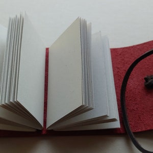 May include: A small red leather-bound journal with a brown leather strap and a white paper interior. The journal is open to reveal the blank pages.
