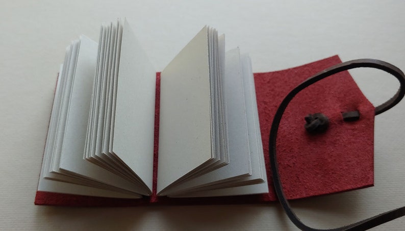 May include: A small red leather-bound journal with a brown leather strap and a white paper interior. The journal is open to reveal the blank pages.