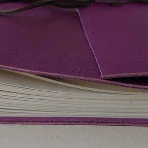 Puede incluir: Un diario encuadernado en cuero morado con un interior de papel blanco. El diario est&aacute; cerrado y atado con una correa de cuero marr&oacute;n.