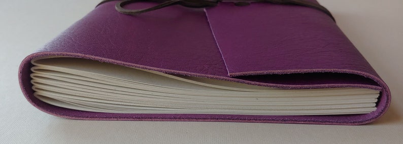 Puede incluir: Un diario encuadernado en cuero morado con un interior de papel blanco. El diario est&aacute; cerrado y atado con una correa de cuero marr&oacute;n.