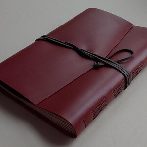 May include: A burgundy leather journal with a wraparound cover and a dark brown leather tie closure. The journal's spine features decorative stitching. The cover is folded over the pages.
