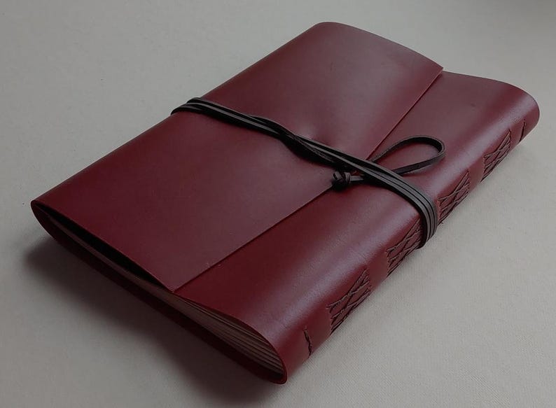 May include: A burgundy leather journal with a wraparound cover and a dark brown leather tie closure. The journal's spine features decorative stitching. The cover is folded over the pages.