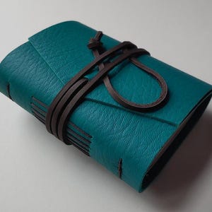 May include: A small, rectangular journal bound in textured teal leather. The cover is secured by a long, thin dark brown leather cord that wraps around the center multiple times and is tied in a simple knot on the front side of the notebook.