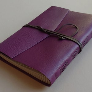 Puede incluir: Un diario encuadernado en cuero morado con una correa de cuero marr&oacute;n y un patr&oacute;n de costura decorativo en la portada.