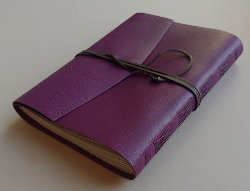 Puede incluir: Un diario encuadernado en cuero morado con una correa de cuero marr&oacute;n y un patr&oacute;n de costura decorativo en la portada.