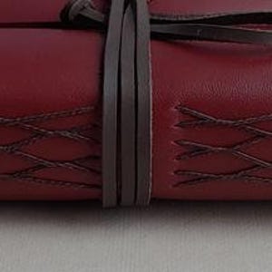 May include: A burgundy leather journal with decorative stitching and a wraparound leather tie. The journal's cover features a textured surface and a dark brown tie to keep it closed. The stitching is a darker shade of brown.